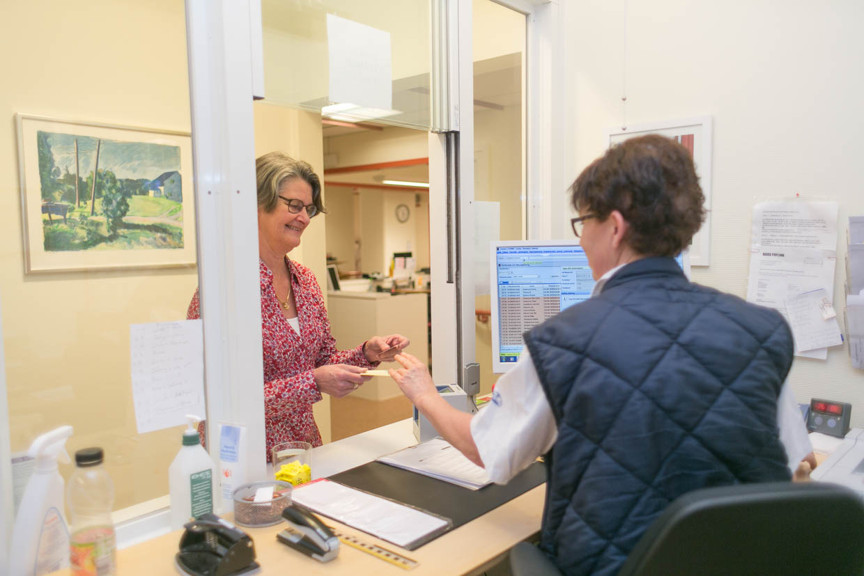 En patient och en vårdpersonal pratar genom en lucka i en reception.