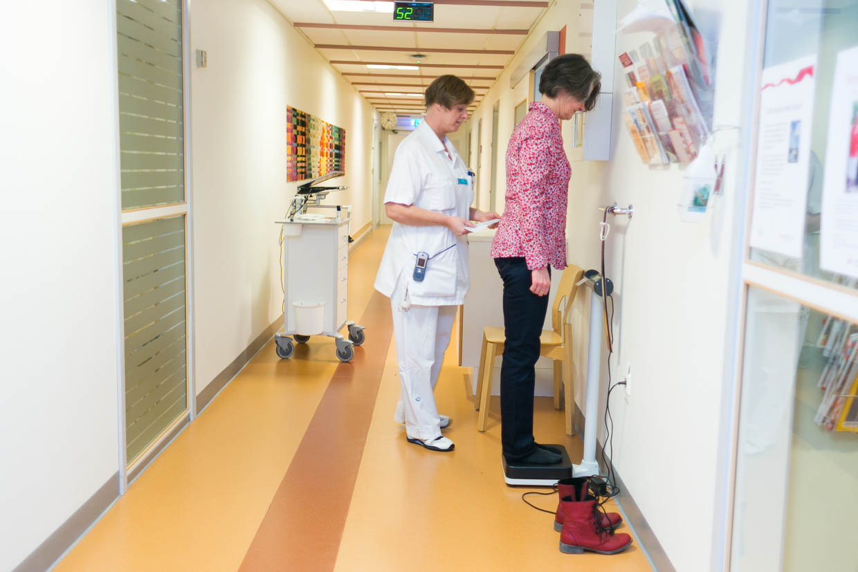 En patient väger sig i en korridor. En vårdperson står bakom och läser på ett papper.