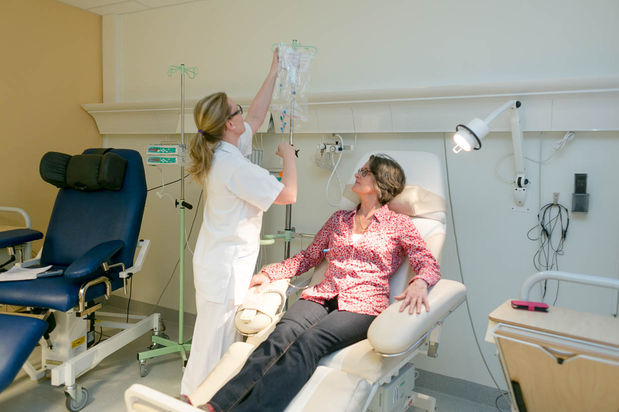 En patient sitter i en fåtölj i ett vårdrum medan en vårdpersonal hänger upp en droppåse i en ställning.