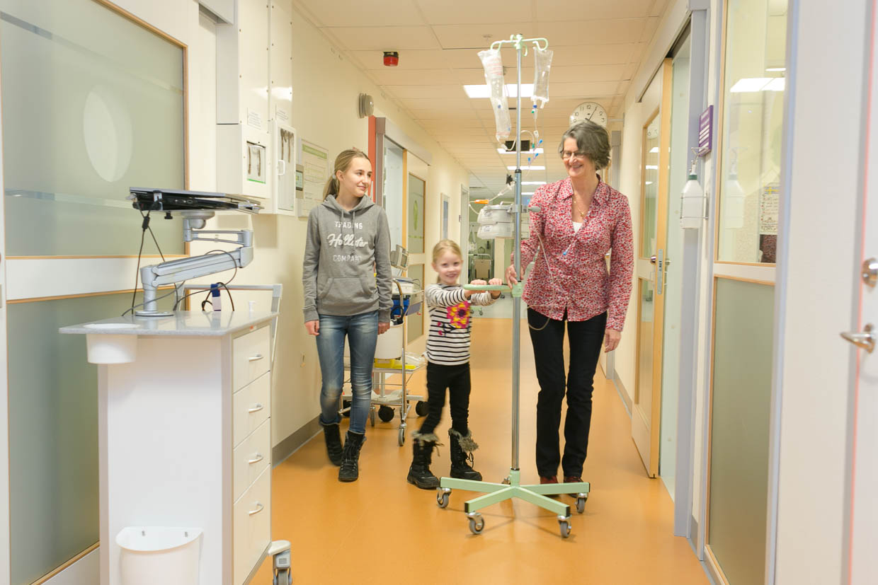 En patient går med en droppställning i en korridor tillsammans med två barn.