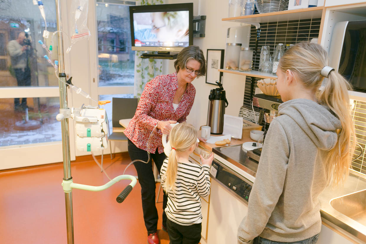 En patient med droppställning häller upp dryck ur en kanna åt ett barn. Ett annat barn står bredvid och tittar på.
