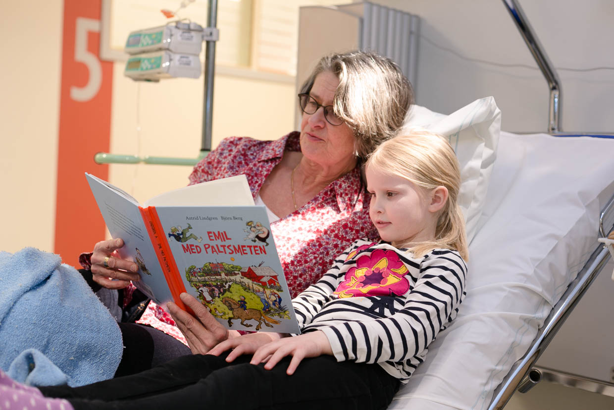 En vuxen och ett barn sitter och läser en barnbok i en vårdsäng.